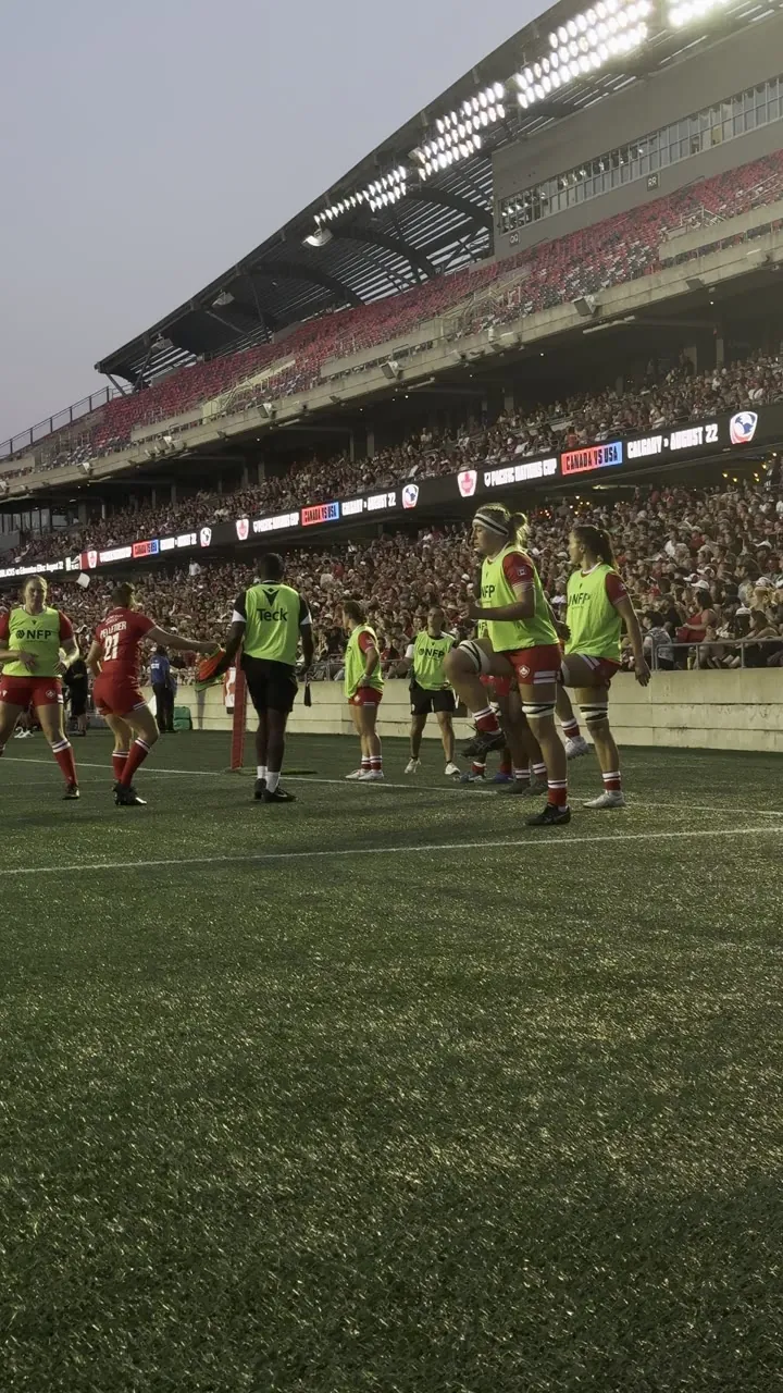 Players in neon bibs on a grass pitch in front of a stadium stand.