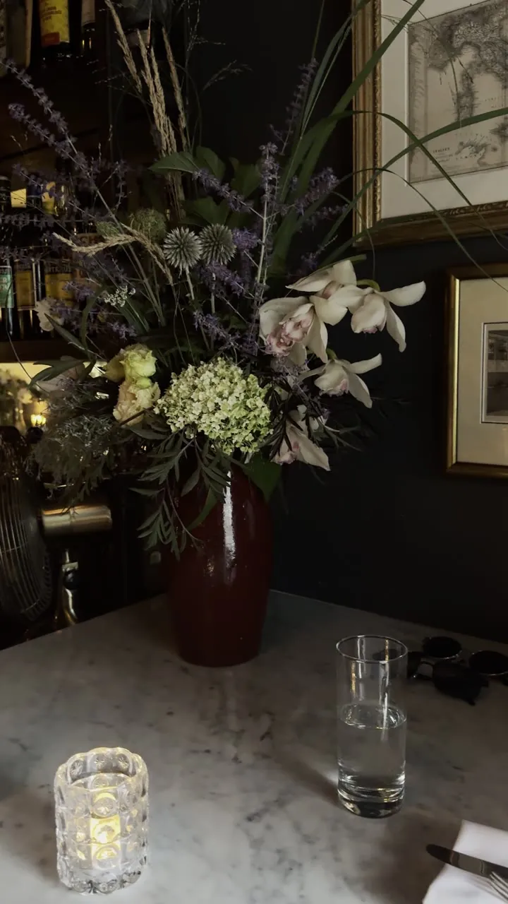 Vase of flowers on a table in a dim restaurant, with a lit candle and a glass.