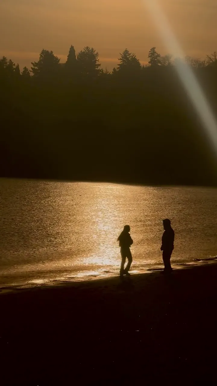Two people silhouetted on a lakeshore at sunset, with dark trees across the water and an orange glow on the surface.
