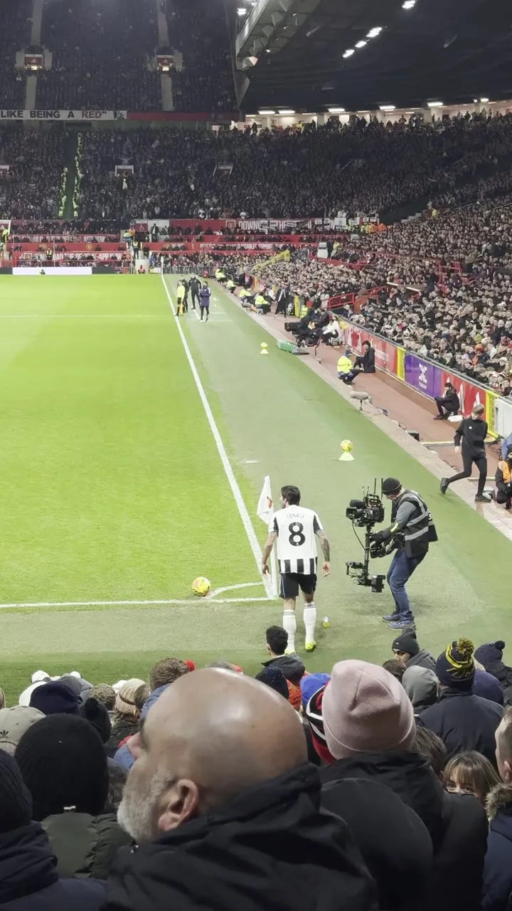 Soccer match seen from the stands, with players on the touchline and spectators in the foreground.