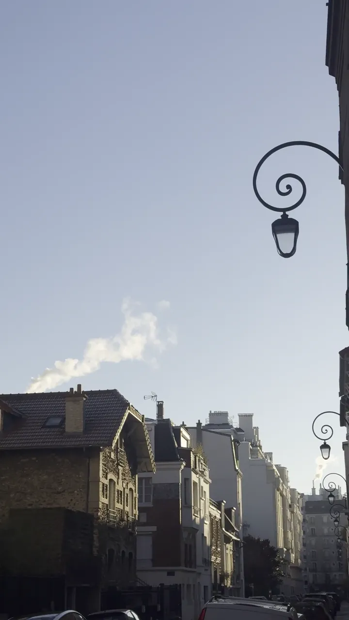 Curled iron street lamp against a pale blue sky, beside stone buildings with a white plume rising behind the roofs.