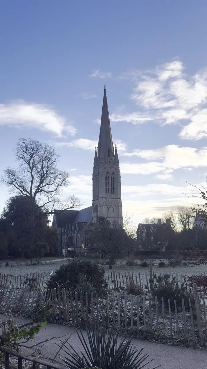 Church spire rising above a snowy garden and bare trees, with clouds in a blue sky.