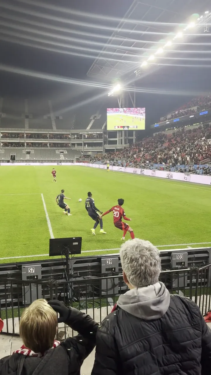 Soccer match seen from the stands, with players on a green pitch under stadium lights and spectators in the foreground.