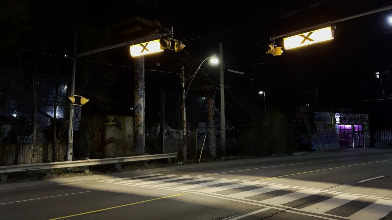 Night street with lit overhead signs and a crosswalk, and a blue car driving through the frame.