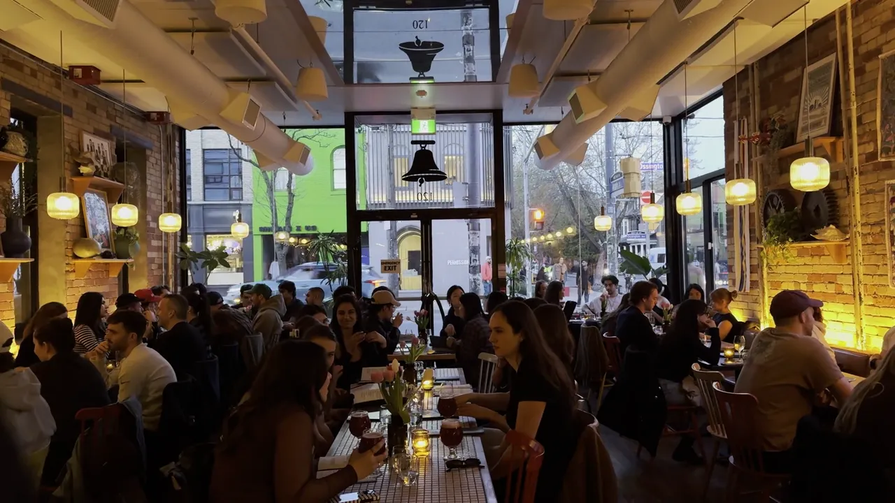 Crowded restaurant interior with warm string lights, diners at long tables and large windows behind.