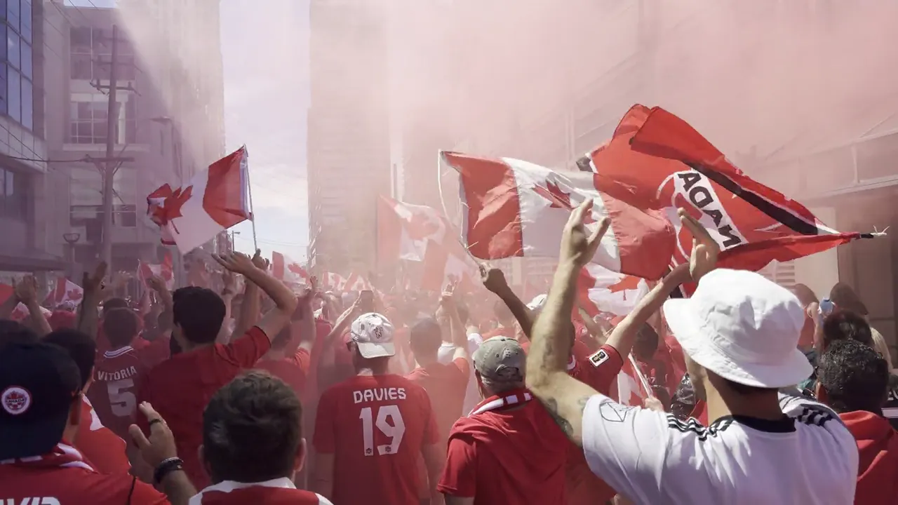 Crowd of supporters in red jerseys waving red and white flags, including Canadian maple leaf flags, through red smoke between buildings.