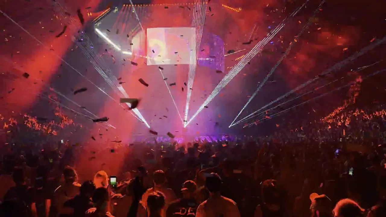 Packed arena crowd facing a stage with a video screen, laser beams and confetti, the lighting shifting from green to red to purple.