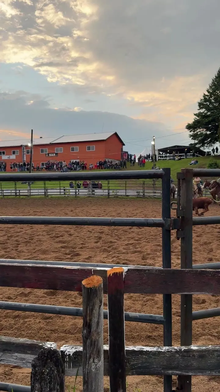 Wooden fence around a dirt riding ring with a red barn and cloudy sky.