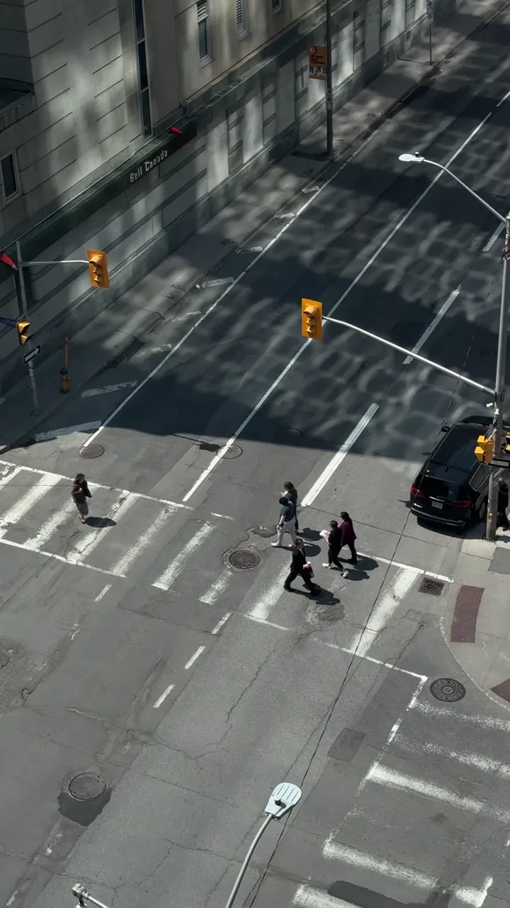 High view looking down on a city intersection, with people crossing at the crosswalks and a dark vehicle waiting.