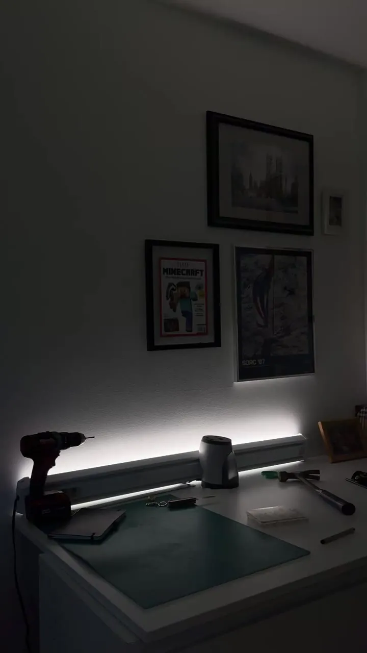 Desk with a lamp under framed pictures in a dim room, lit by a white strip light.