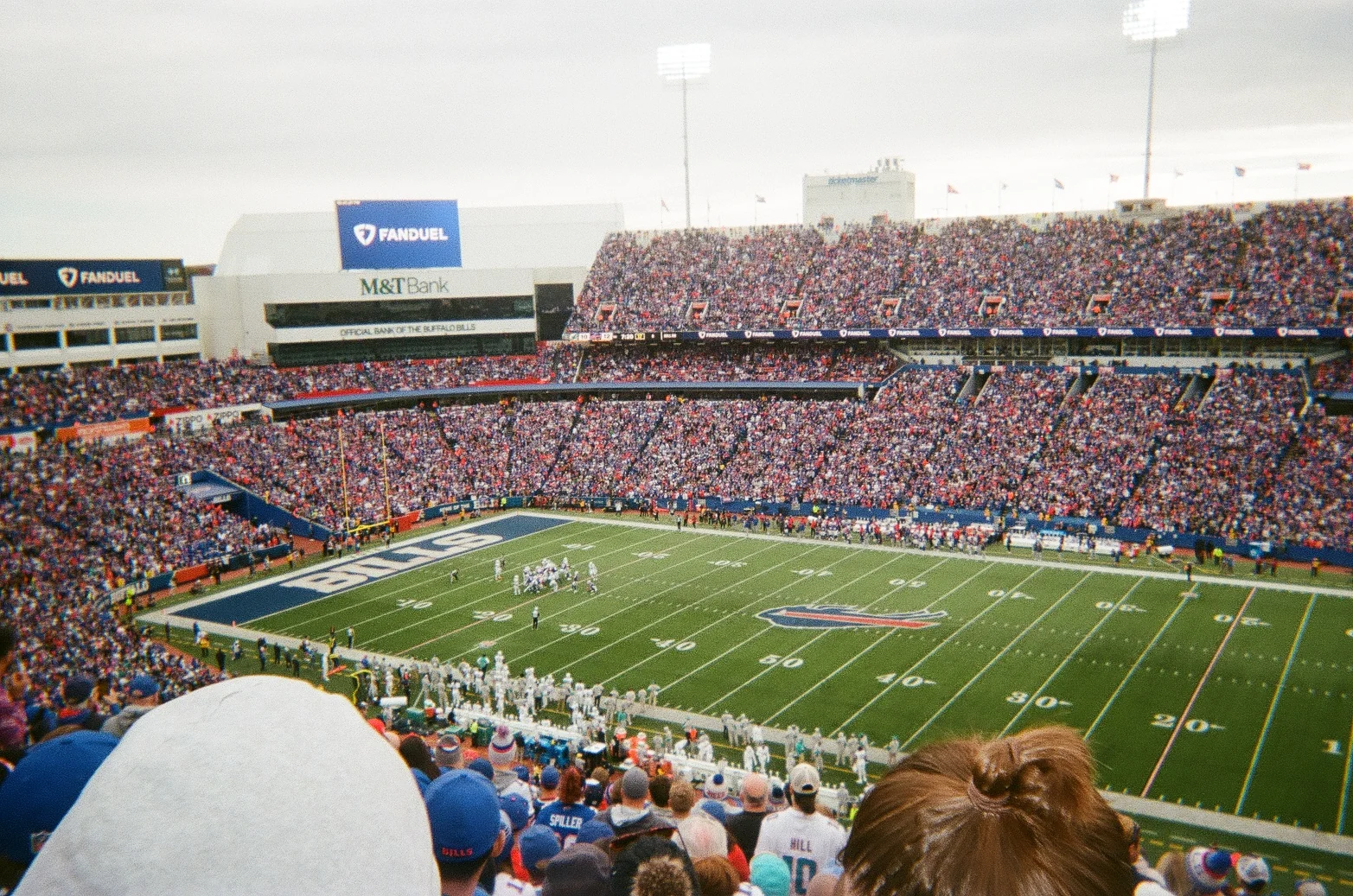 A view across a large football stadium full of spectators, with the green field and yard lines below, a tall sign above the stands and the back of a person's head and a grey cap in the foreground.
