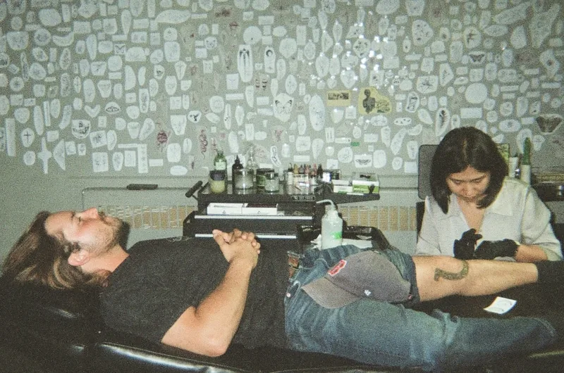 A man with long hair and a beard lies back on a black chair while a person in a white shirt and black gloves works on a tattoo on his leg, in front of a wall covered in stickers.