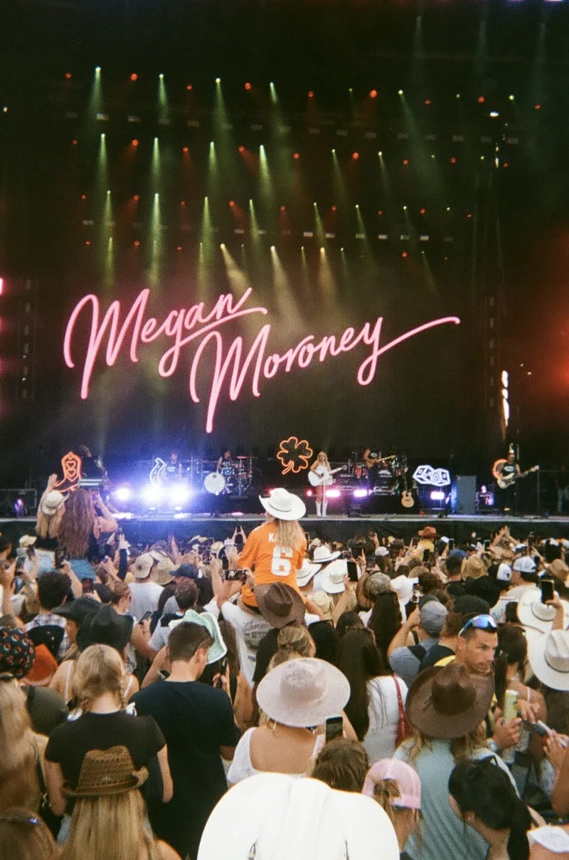 A crowd watches a stage with bright overhead lights and a pink neon sign reading Megan Moroney, with a few people in cowboy hats in the foreground.