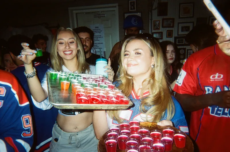 Two smiling young women with long blonde hair hold a tray of red and green shot cups at a crowded party, with men in hockey jerseys and a person in a cap behind them.