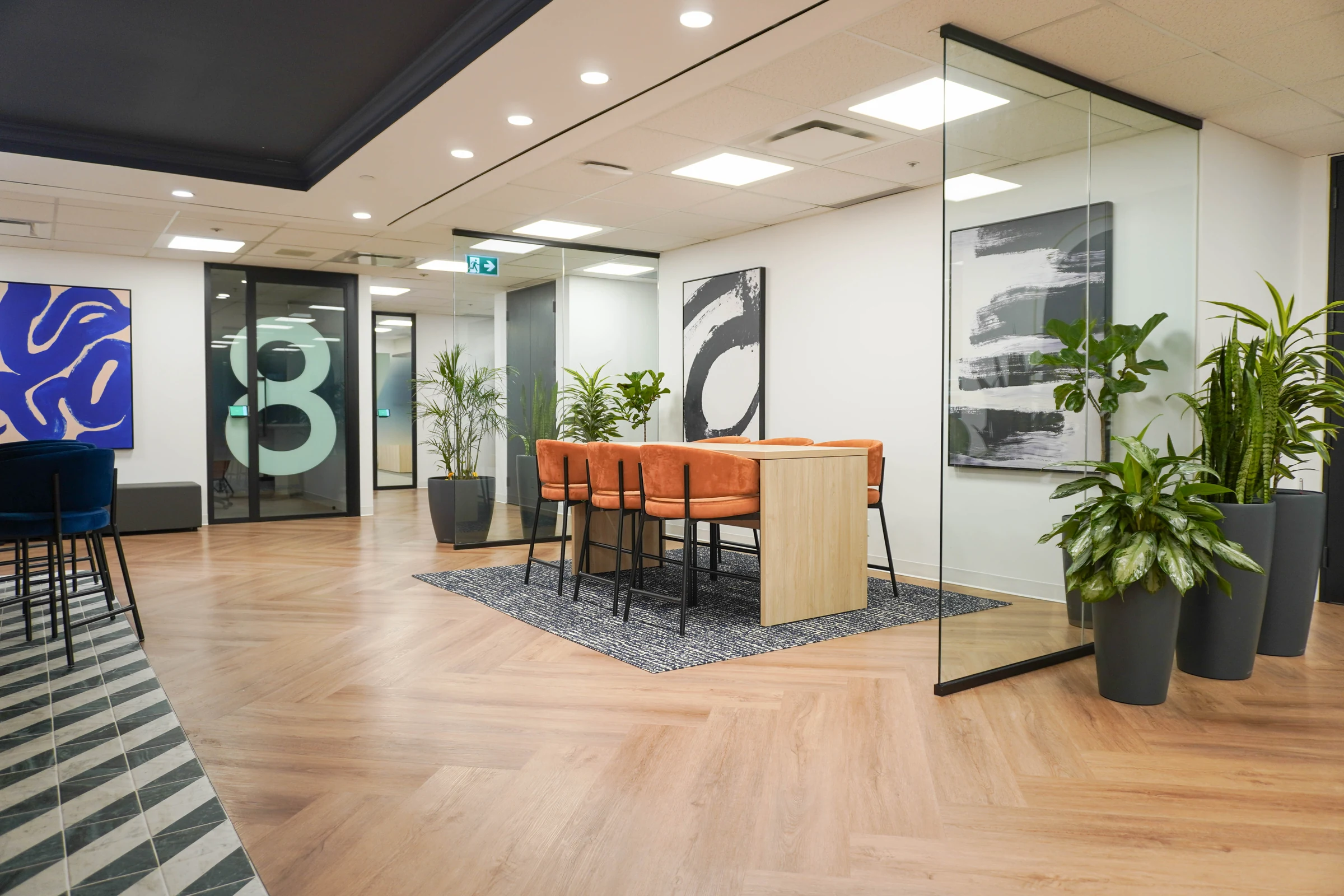Open reception area with a herringbone wood floor, a high table with orange stools on a patterned rug, large framed abstract artworks, potted plants and glass partitions, one with a large number 8.