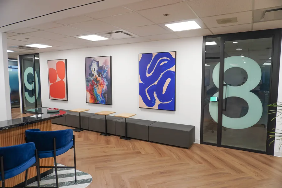 Entry hallway with three framed colourful abstract artworks on a white wall, grey bench seating, two blue chairs in the foreground and a glass door with a large pale number 8.