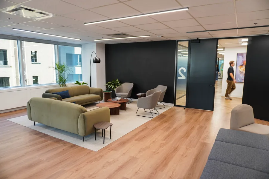 Open lounge with olive-green sofas, grey armchairs and low tables on a cream rug, a dark partition wall and large windows; a person stands in the background.