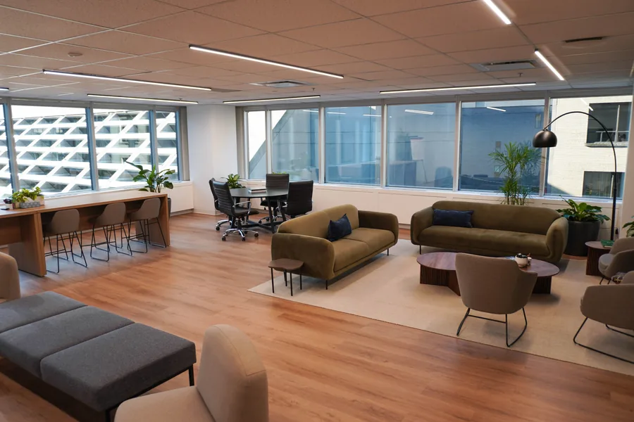 Lounge by tall windows with olive sofas, a low table and a dark chair, a high counter and meeting table beyond, and a grey cushioned bench in the foreground.