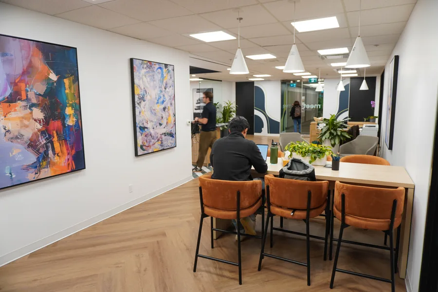 Kitchen seating area with orange chairs at a counter, two framed colourful abstract paintings on a white wall and pendant lights; two people are at the far end.