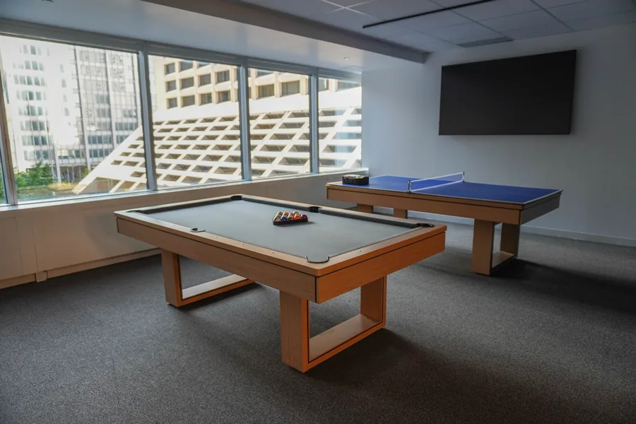 Games room with a pool table and a ping-pong table on grey carpet, a wall-mounted TV and tall windows facing nearby buildings.