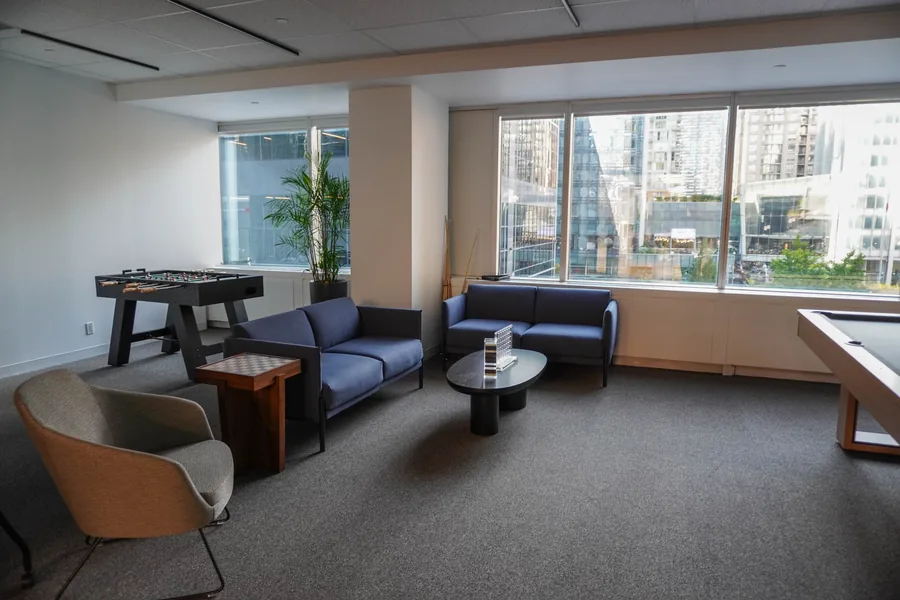 Games room lounge with blue sofas, a small game table, wooden side tables and a tan armchair beside large windows; the edge of a pool table is visible at right.