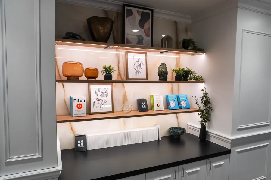 Built-in white shelving niche with warm lighting, holding books, a framed print, pots and small objects above a black countertop.