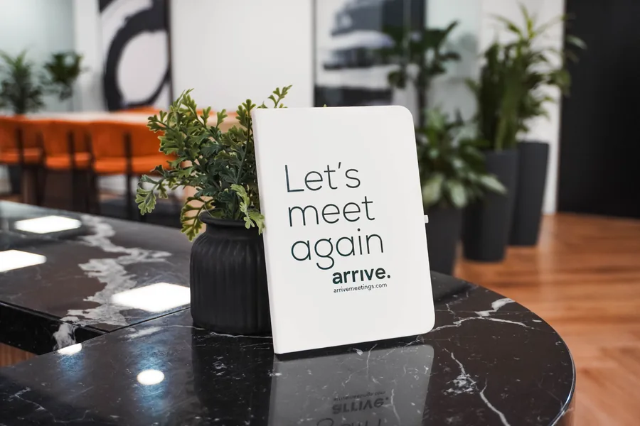 A white card reading Let's meet again, with a small wordmark beneath, standing on a black marble tabletop beside a potted plant, with orange chairs blurred behind.