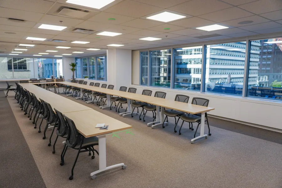 Long boardroom with two lines of light wood tables and black mesh chairs under rows of ceiling lights, beside a wall of windows.