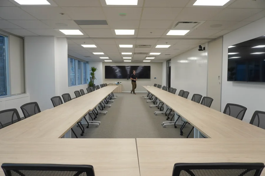 Looking along a large U-shaped boardroom table with black chairs toward a wall-mounted screen; a person stands at the far end.