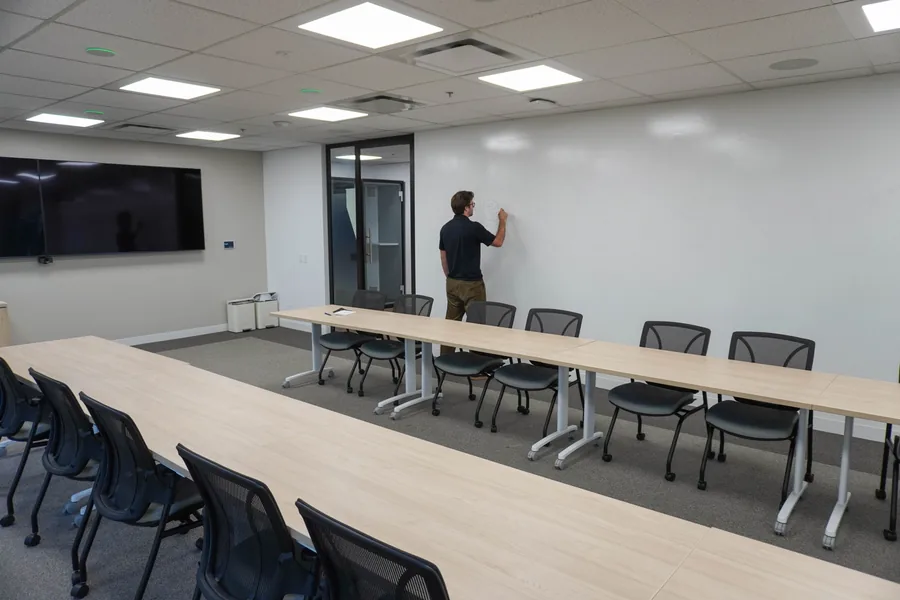 Boardroom with long tables and black chairs; a person at the back wall writes on the white wall beside a wall-mounted screen.