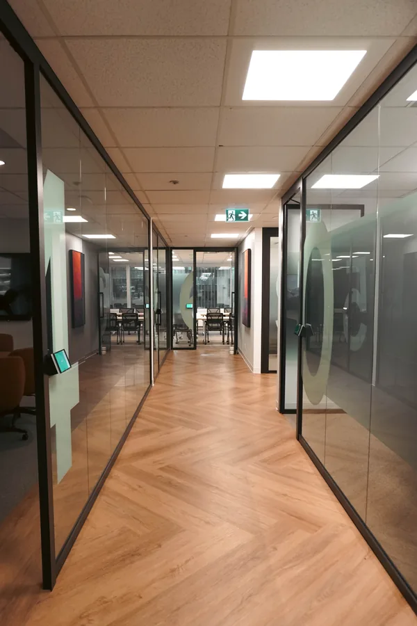 Corridor with glass-walled offices on both sides, a herringbone wood floor and ceiling panel lights.