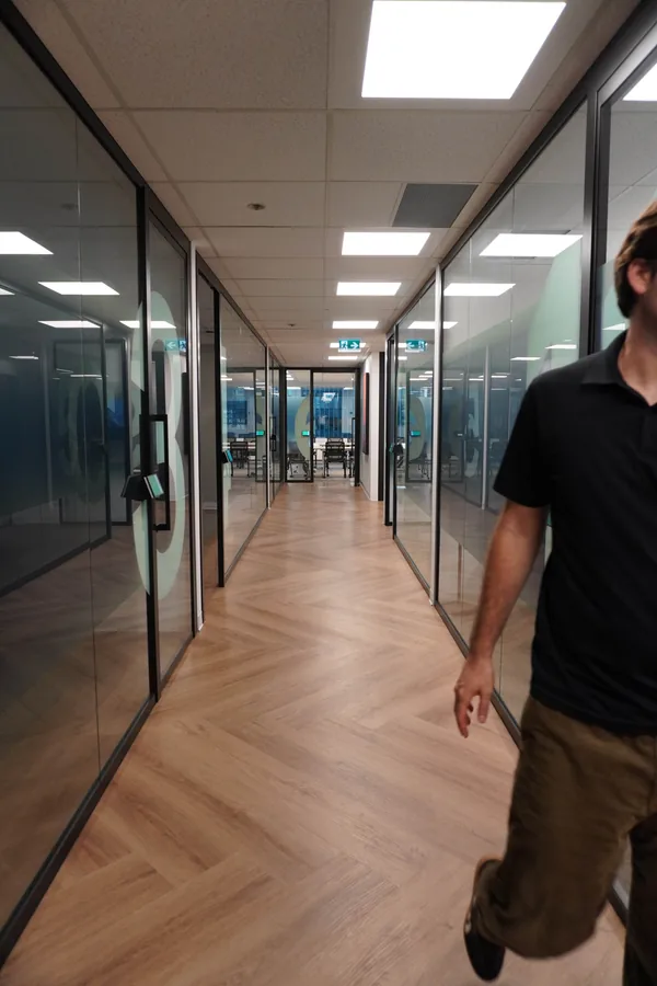 Glass-walled corridor with a herringbone floor; a person in a black shirt walks at the right edge of the frame, partly out of shot.