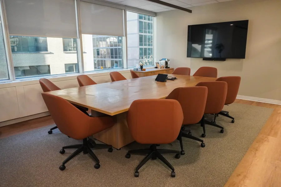Meeting room with a wooden oval table and orange chairs, a wall-mounted TV and window blinds, on a grey rug.