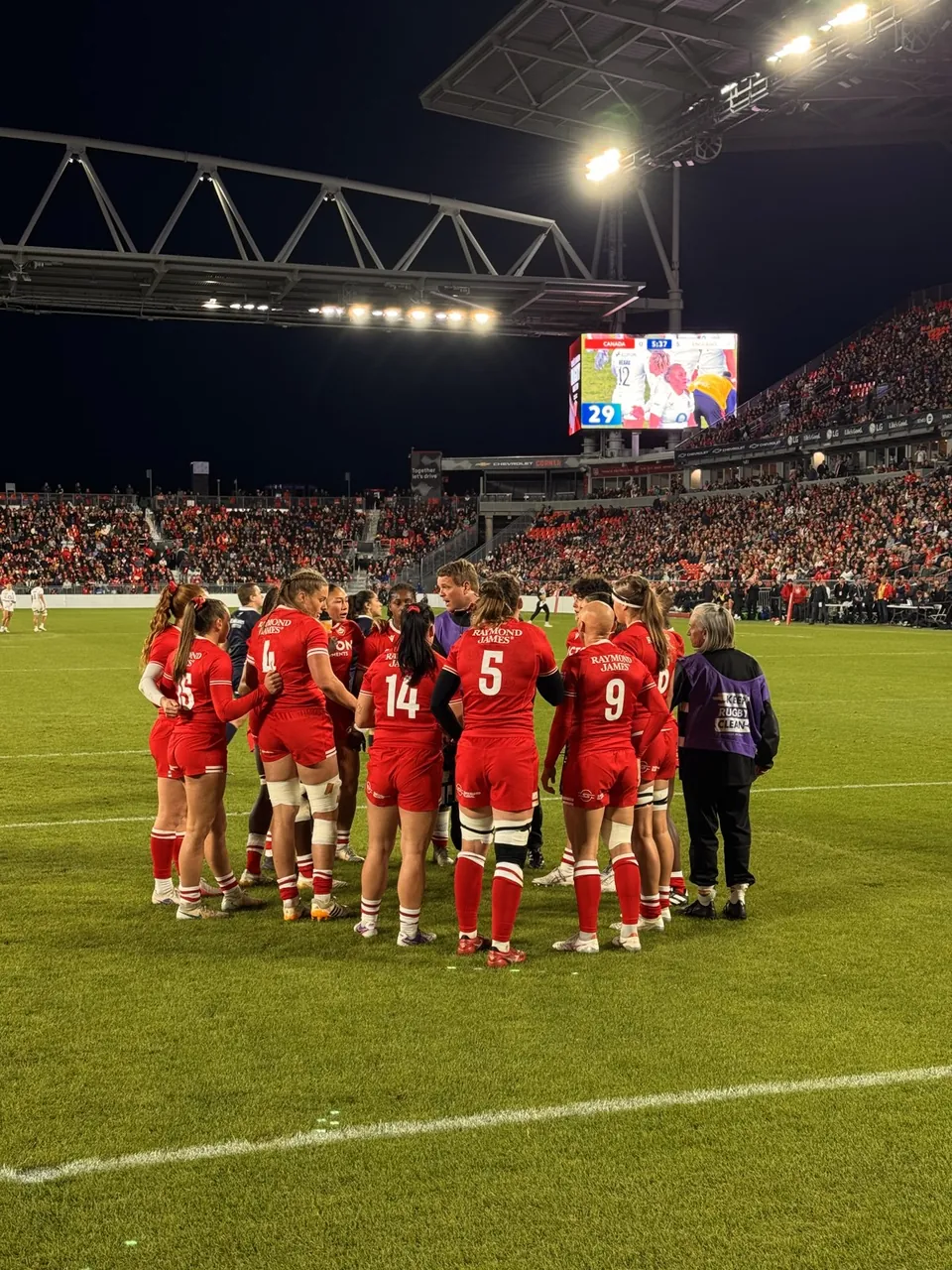 Canada women’s team in a huddle on the pitch with the Raymond James content on the jumbotron behind.