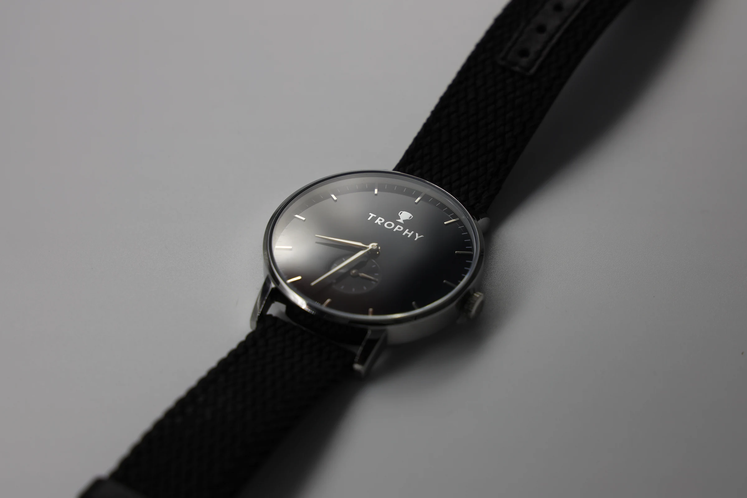 A wristwatch with a black dial, silver case and woven black strap lies at an angle on a grey surface; the word TROPHY is printed on the dial.