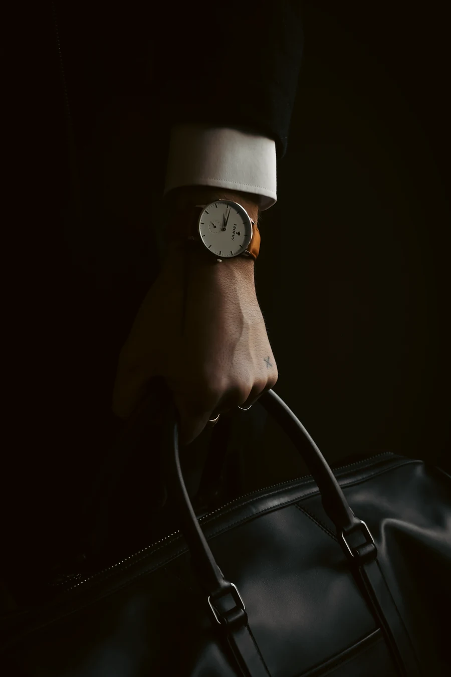 A hand in a dark sleeve with a white cuff, wearing a watch with a white dial and tan strap, holds the handles of a black leather bag in a very dark scene.