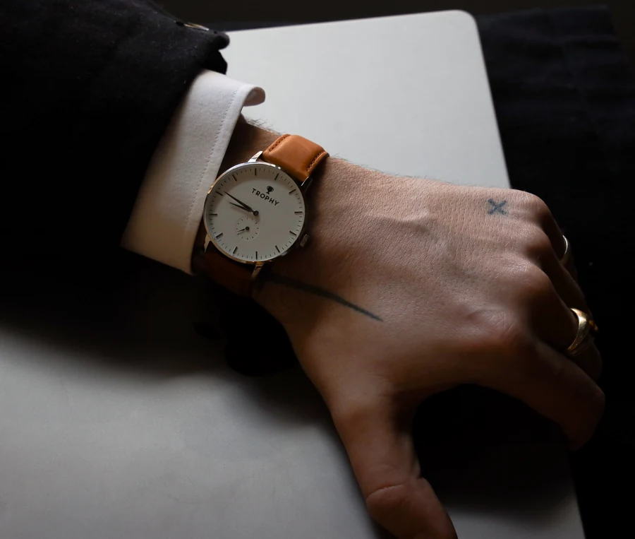 A wrist in a dark jacket with a white shirt cuff wears a watch with a white dial and tan leather strap, resting on a grey laptop; the hand has a small cross tattoo and a gold ring.