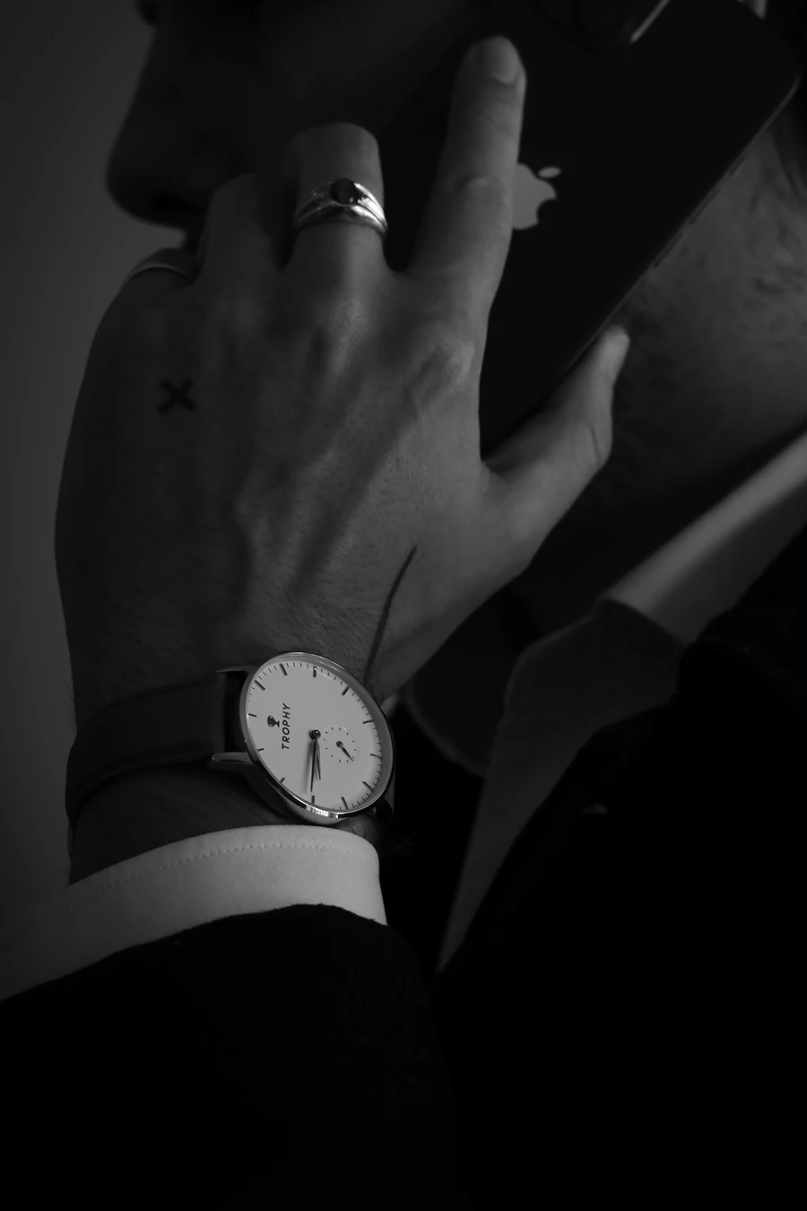 In black and white, a hand with rings and a small cross tattoo holds a phone to a face, with a watch with a white dial on the wrist, a white shirt cuff and a dark suit sleeve.