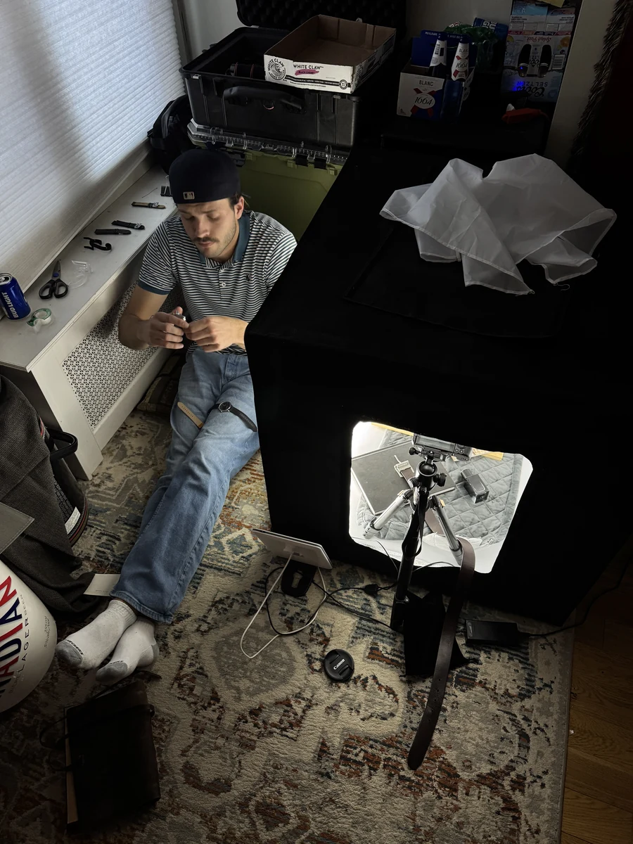 A man in a cap and striped shirt sits on the floor of a room beside a black light box with a bright opening in the front, where a camera on a tripod points inside; crumpled white fabric rests on top, and the floor has a patterned rug.