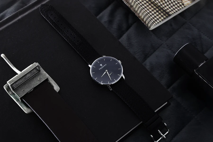 A watch with a dark dial and black strap lies on a black notebook on dark quilted fabric, next to a silver buckle and a black strap, with a patterned fabric corner and a black cylinder at the edges of the frame.