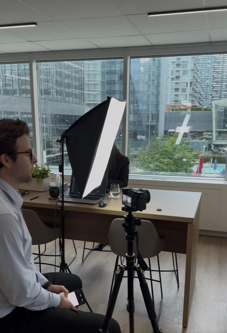 Behind-the-scenes view of the interview setup: a camera on a tripod and a softbox light beside a desk, with a man seated in the foreground.