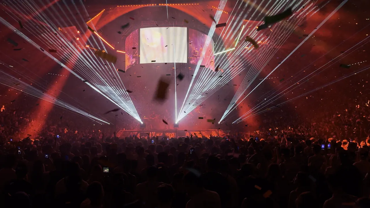Packed arena crowd facing a stage with a video screen, red lighting, laser beams and confetti.