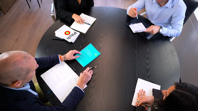 Overhead view of four people seated around a round dark table with notebooks, papers and a teal booklet.