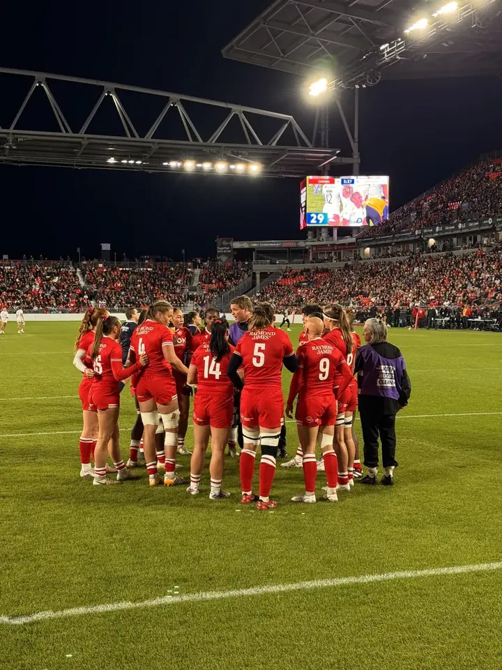 Canada women’s team in a huddle on the pitch, with a lit jumbotron and stadium stands behind.