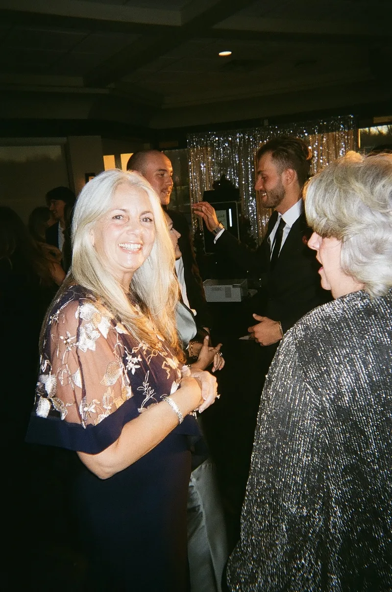 A woman in a sequined dark dress smiles in the foreground as guests in dark clothing talk behind her, with a woman with grey hair beside her.