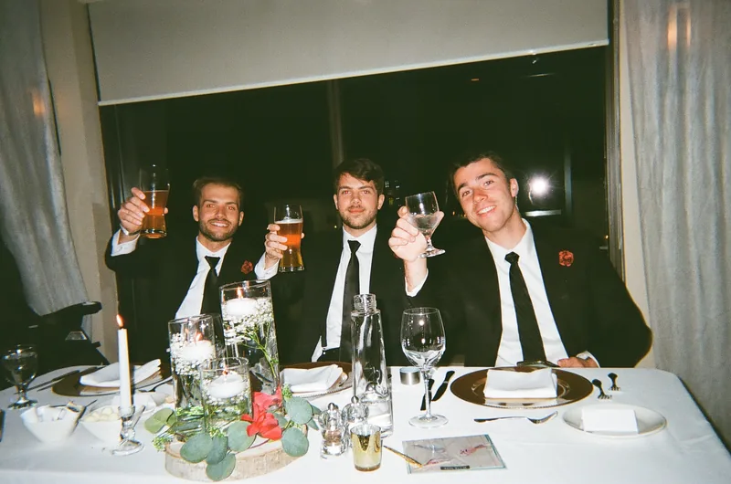 Three men in dark suits and ties at a table raise drinks towards the camera, with candles, flowers and glasses in the foreground.