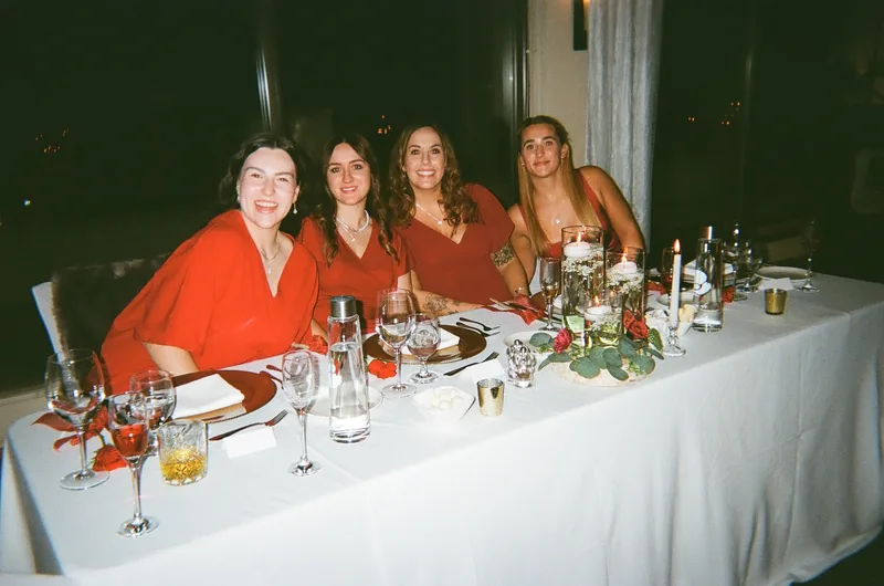 Four women in red and orange dresses smile from behind a long white-clothed table set with glasses, candles and flowers, with dark windows behind them.