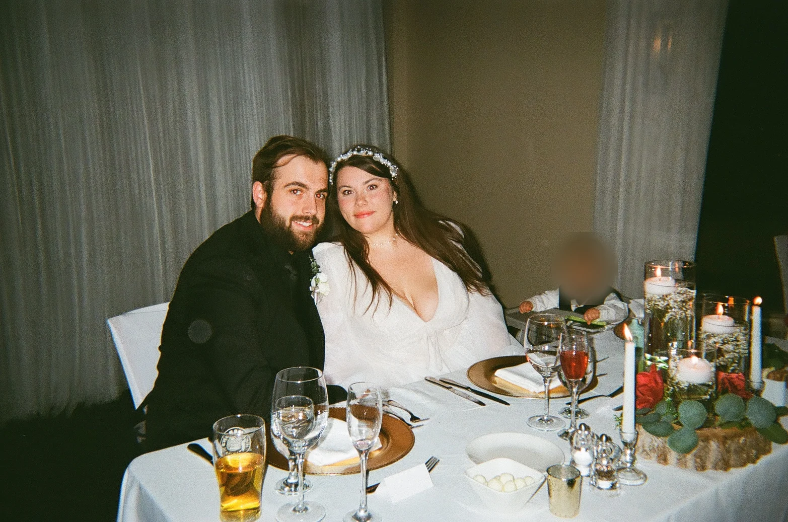 A bearded man in a dark suit and a woman in a white dress with a tiara sit side by side at a white-clothed table with candles, flowers and drinks; a small child in a dark vest, with a blurred face, sits behind them.