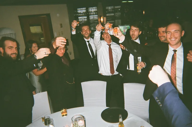 A group of men in suits and ties raise shot glasses and drinks together over a table, with a blue suit sleeve and a raised fist in the foreground.