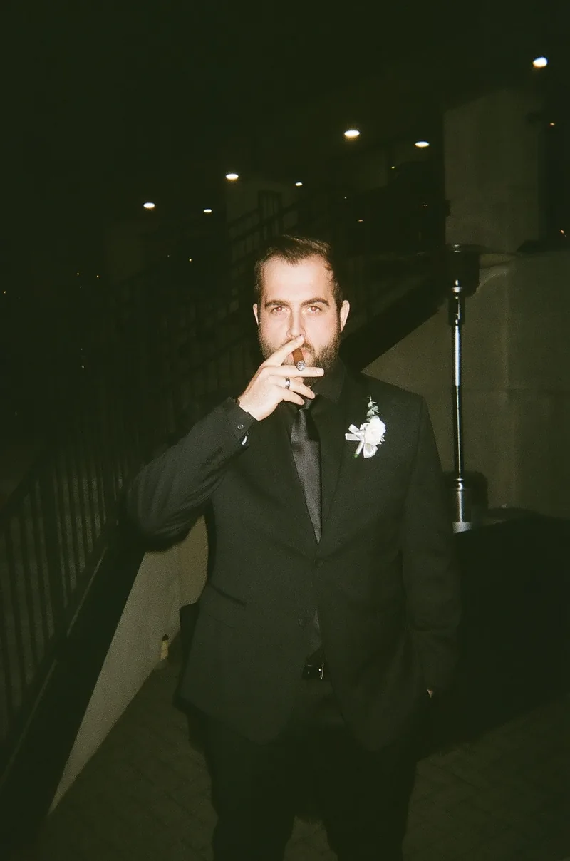 A man in a dark suit with a flower on the lapel holds a cigar to his mouth, standing on a dark terrace at night.